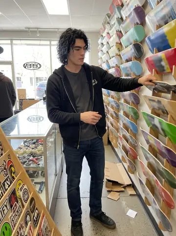 Bryson, AI character: inside the Portland skate shop, between the deck wall and a glass counter of wheels and bearings