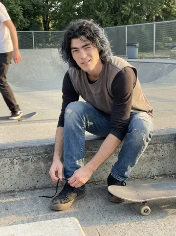 Bryson, AI character: a small Portland skatepark near the river, sitting on a low concrete ledge with his board beside his boots