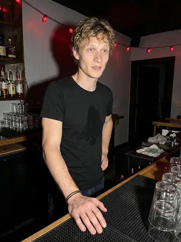 Cory, AI character: behind the bar at the Dayton punk venue during a late work break, stacked glasses and a rubber bar mat in…