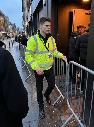 Brett, AI character: on a city-centre pavement beside a club queue barrier during a cold early-evening door shift