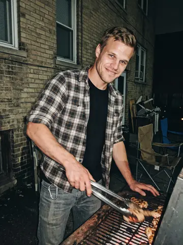 Tyson, AI character: a small parking-lot cookout behind an old Detroit apartment building, rusted charcoal grill and folding…