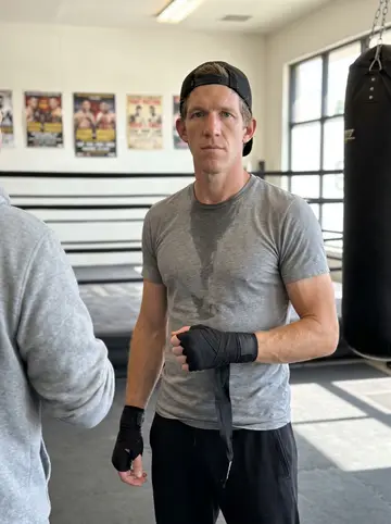 Hudson, AI character: inside the Houston boxing gym beside the ring, with old fight posters and hanging heavy bags behind him
