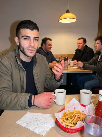 Strahinja, AI character: a quiet Tuesday-night diner booth in Calgary, coffee mugs, fries, and a folded paper napkin with a chess…