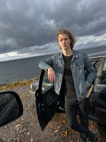 Leevi, AI character: a windy turnout above Lake Superior outside Duluth, his old dark sedan parked on wet gravel with storm…
