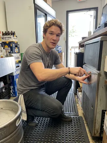 Gijs, AI character: behind the bar during a slow afternoon, crouched beside the open ice machine with beer cases and a rubber…