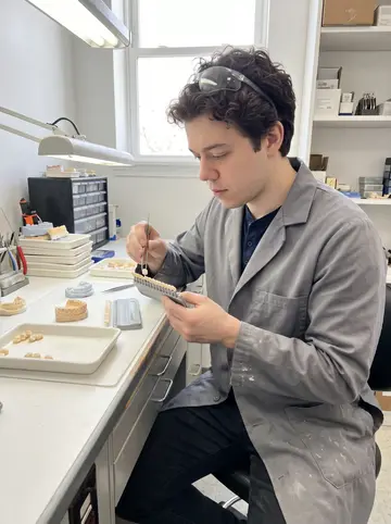 Kostiantyn, AI character: a small dental lab in Philadelphia, standing at a cluttered workbench with ceramic trays, molds, and a…