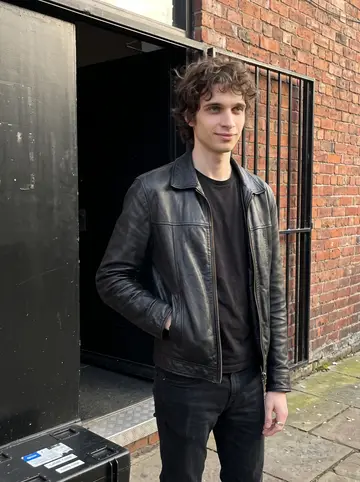 Finbar, AI character: outside a Liverpool venue stage door after afternoon soundcheck, brick wall and black door behind him