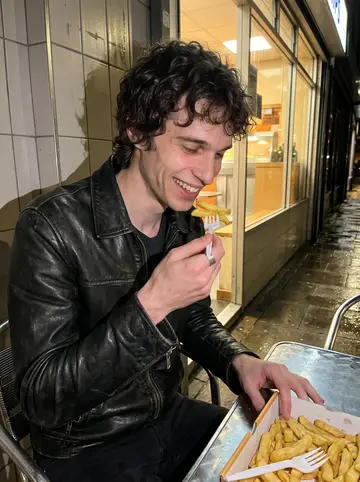 Finbar, AI character: outside a late-night fish and chips shop after a gig, pavement damp and takeaway boxes on a metal table