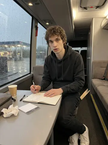 Finbar, AI character: tour bus lounge parked at an M6 service station, rain on the windows, a notebook open on the table