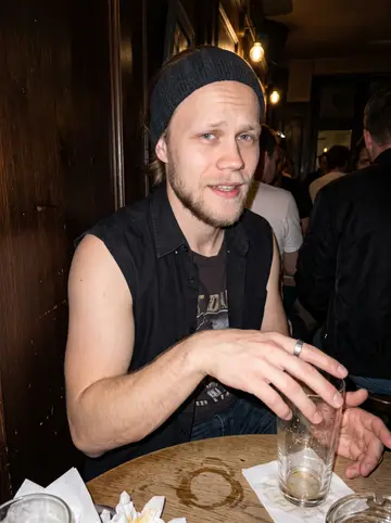 Verneri, AI character: a crowded little bar near the venue in Cologne after a gig, dark wood wall and half-empty glasses behind him