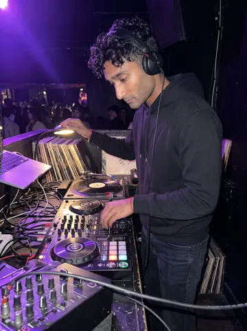 Tejas, AI character: DJ booth in a narrow East Sixth club during his late slot, mixer and turntables crowded into the corner