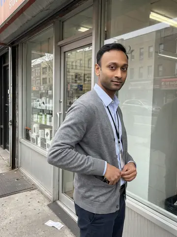 Akash, AI character: outside his small phone repair shop on a bright spring afternoon in Jackson Heights, leaning against the…
