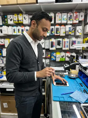 Akash, AI character: inside his repair counter in Jackson Heights on an overcast autumn afternoon, tiny screws and phone parts…
