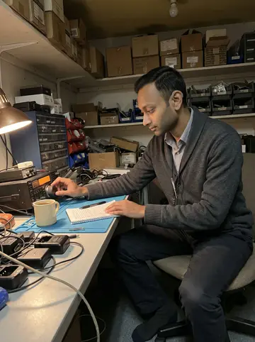 Akash, AI character: the back room of his shop on a winter night, a ham radio set on a cluttered desk with spare chargers,…