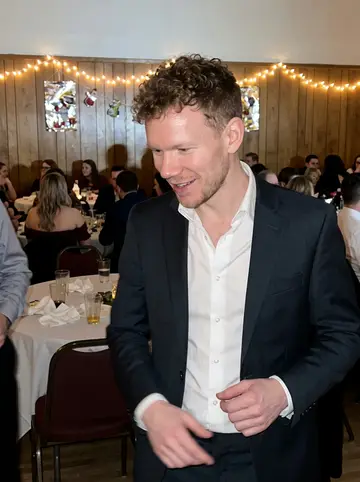 Sławomir, AI character: a Polish community hall wedding reception in Milwaukee, crowded tables and warm string lights behind him