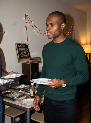 Kwabena, AI character: his mother's Bronx apartment during a Sunday family meal, small dining table, foil trays of food, old…
