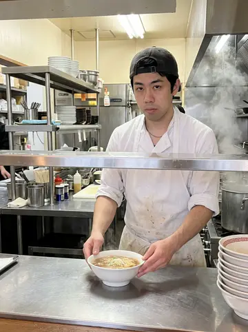 Shota, AI character: early morning prep area inside the noodle shop before opening, stock pots, metal counter, stacked bowls…