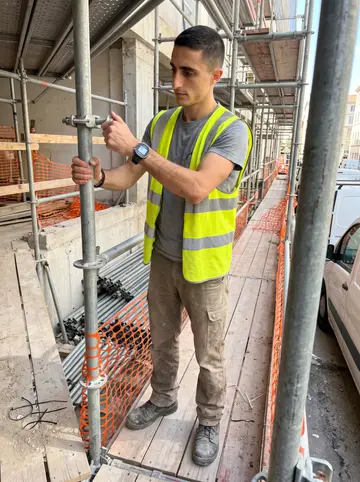 Cédric, AI character: a scaffold frame beside a renovation site near La Joliette, stacked tubes and orange netting around him