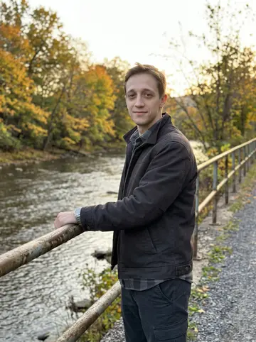 Dale, AI character: the Lackawanna River Heritage Trail in Scranton, leaning against a weathered metal railing with autumn…