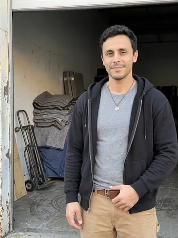Eliseo, AI character: the open loading bay of a small South Philly moving warehouse after a job, stacked moving blankets and a…