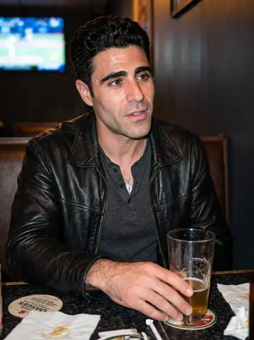 Rodolfo, AI character: a neighborhood bar on Atwells Avenue on a humid summer night, sitting with one friend while a baseball…