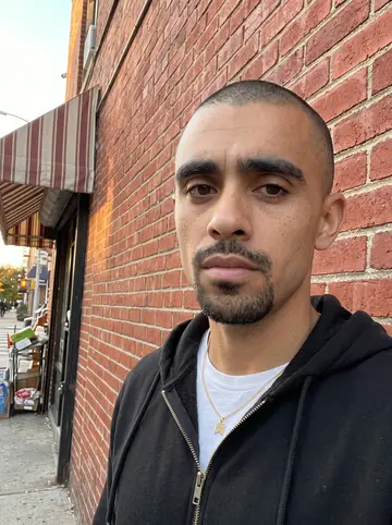 Luis, AI character: on the corner outside his abuela's bodega in North Philly, leaning on the red-brick wall beside the door,…