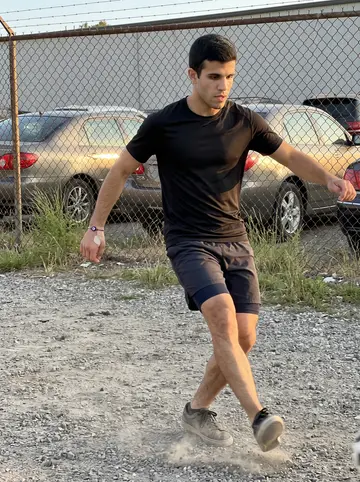 Damianos, AI character: a gravel patch behind the warehouse used for pickup soccer, chain-link fence, parked cars and scrub grass…