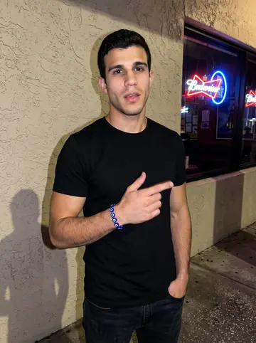 Damianos, AI character: outside a small Tarpon Springs bar after a cousin's birthday, stucco wall, neon beer sign in the window,…