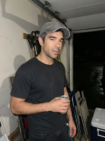 Mário, AI character: a cousin's open garage after a Sox game on the radio, folding chairs and a cooler behind him, damp…