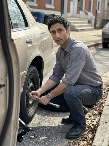 Aidan, AI character: curbside on a Baltimore rowhouse block, helping change a flat tire on an older sedan
