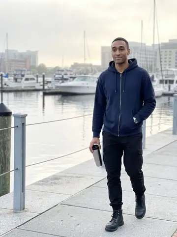 Obinna, AI character: the Inner Harbor promenade at 6am, water and moored boats behind him, city buildings faint in morning fog