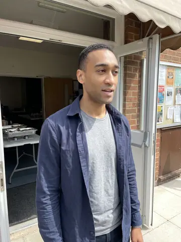 Obinna, AI character: outside his mother's church hall after first-Sunday lunch, folding tables and foil trays just visible…