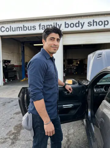 Ravi, AI character: outside the Columbus family body shop after closing, beside the restored 1967 Mustang with the shop doors…