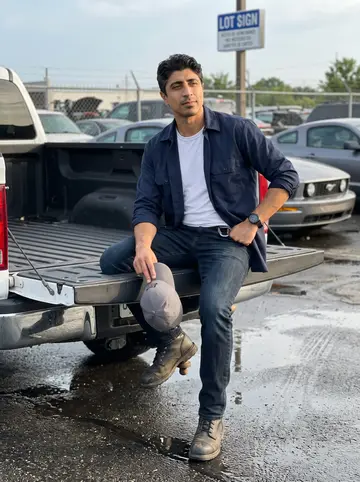 Ravi, AI character: a wet parking lot outside a parts yard on the edge of Columbus, the Mustang parked behind him with rain…