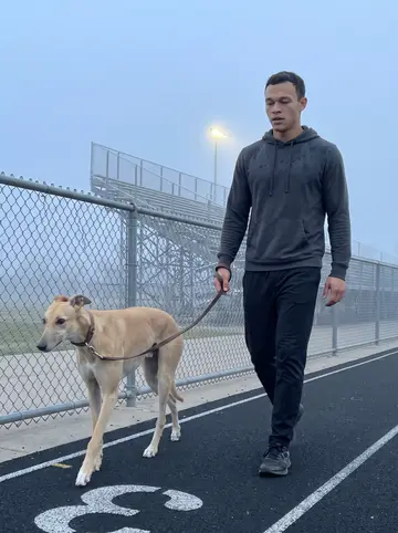 Hunter, AI character: the empty high-school track before dawn in Tulsa, chain-link fence and bleachers fading into light fog