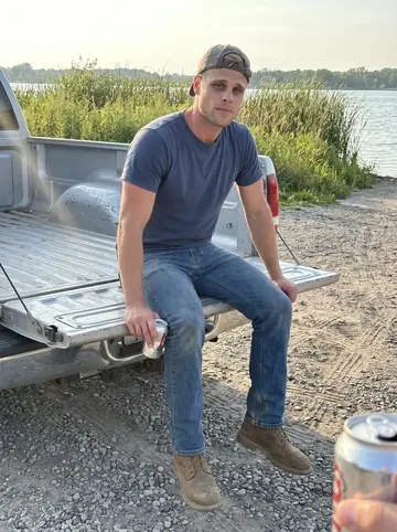 Duncan, AI character: gravel lot by the lake launch, his worn silver 2011 F-150 with a dented tailgate, tall grass behind