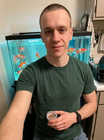 Jared, AI character: his small Akron apartment beside the rectangular aquarium, seven orange-and-white goldfish visible in the…