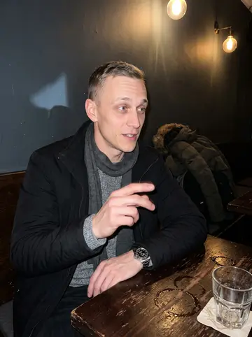 Michał, AI character: a small Hamburg Kneipe after work, dark wood table, coats piled on the bench beside him