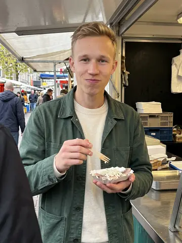 Bas, AI character: a Saturday market stall near Binnenrotte, standing by a small counter with herring and onions