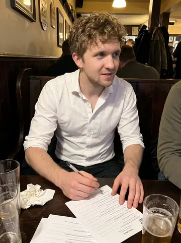 Eoin, AI character: a crowded Dorchester pub quiz table with pint glasses, trivia sheets, and dark wood booths around him