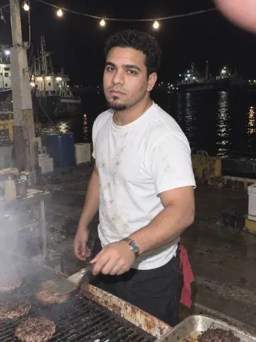 Marcos, AI character: on the marina dock at night, a rusty grill and string lights, dock hands waiting for plates