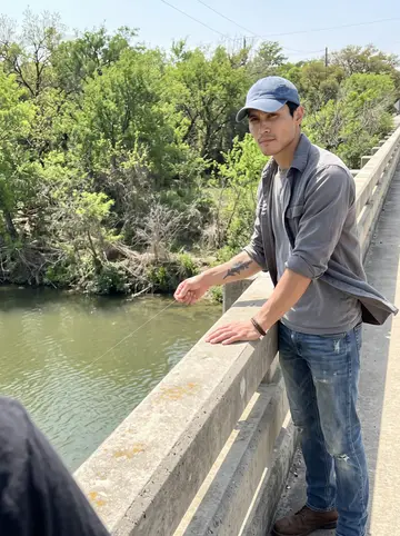 Diarmuid, AI character: a concrete bridge over the Medina River in bright spring sun, fishing line dropped from the rail and…