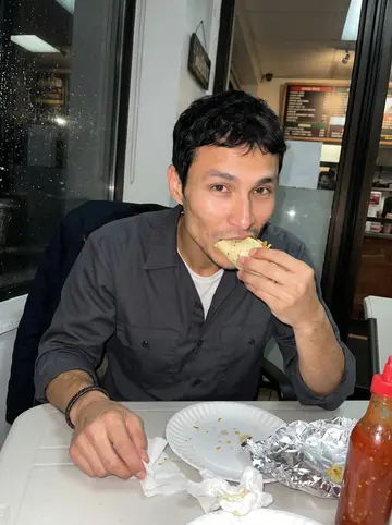 Diarmuid, AI character: inside a small late-night taco shop near his apartment, plastic table, red salsa bottle, and rain-dark…