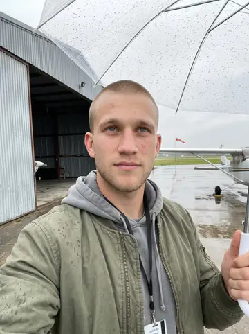 Barrett, AI character: the airfield ramp above Stonington, Maine, leaning against the wheel strut of a single-engine plane,…