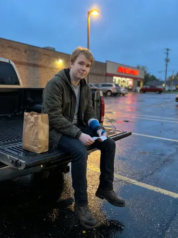 Kingston, AI character: a damp parking lot outside an auto parts store near Milwaukee, his old pickup parked under a sodium…