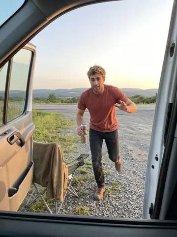 Freddie, AI character: a Finger Lakes roadside pull-off at sunset, his lived-in van parked with the side door open and a folding…