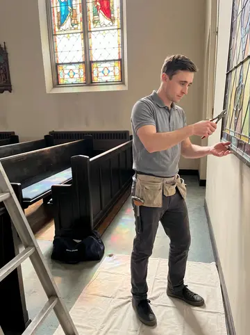 Dawid, AI character: inside an old Buffalo church while he repairs a stained glass panel, ladder and protective cloth on the floor