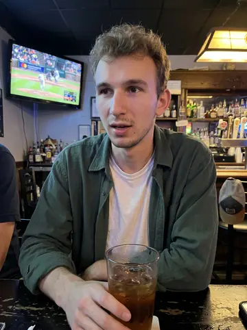 Krzysztof, AI character: a small neighborhood bar in Detroit with a Tigers game playing on a wall TV and friends cropped around the…
