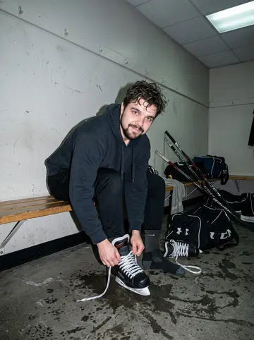 Jovan, AI character: a community rink locker area after Thursday pickup hockey, concrete floor wet near the benches, hockey…