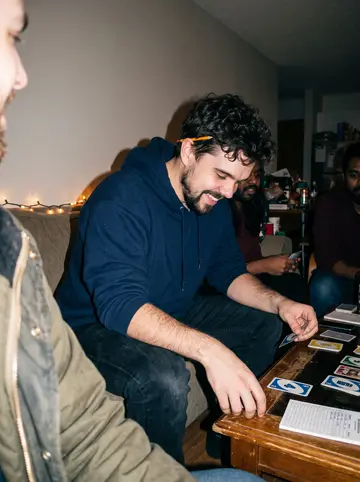 Jovan, AI character: a crowded apartment living room during a low-key holiday game night, card game on the coffee table and…